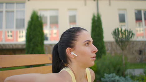 A-young-woman-with-headphones-performs-push-ups-on-a-bench-in-a-city-park-in-slow-motion.-Training-a-young-woman-on-a-bench-against-the-backdrop-of-the-city-and-houses